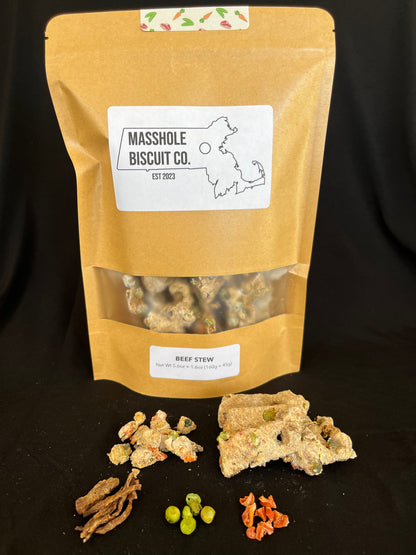 Masshole Biscuit Co all natural dog treat combo bag. Massachusetts state shaped treats with Masshole Biscuit Co training dot shaped treats. Both treats are featured along side USDA beef, peas, and carrots, which serve as the chunky style add ins to the dog treat dough. The Masshole Dog treat package is beige featuring the Masshole Biscuit Co logo and the dog treat flavor, beef stew. Their is a clear window where you can see the Massachusetts state shaped treats.