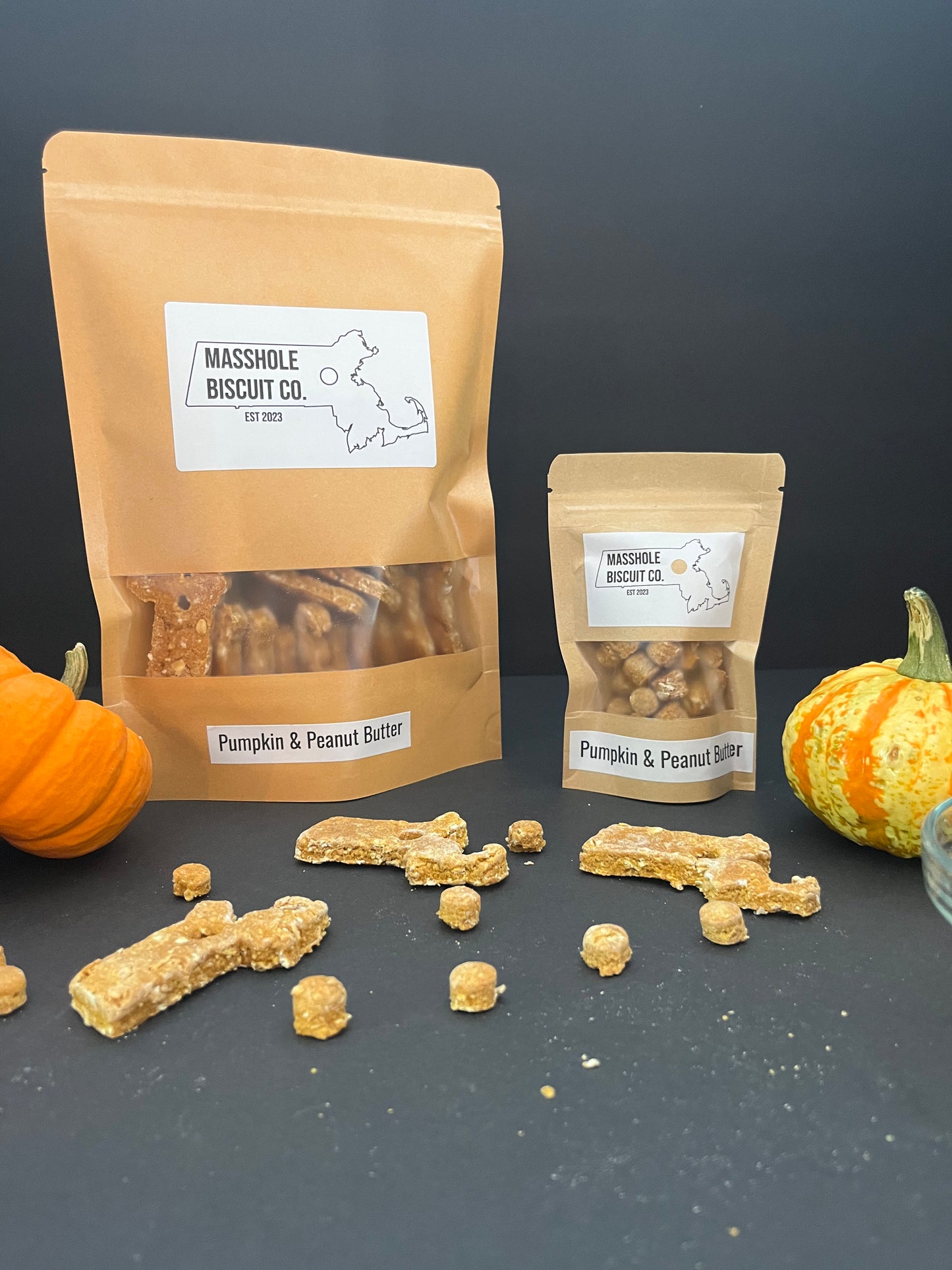 Homemade all natural dog treats made with whole ingredients like pumpkin and peanut butter. Treats are shaped like Massachusetts with holes in the middle. The holes are repurposed to make Masshole dog treats. This picture shows the packaging and the treats. Dog cookie, dog treat, dog snack. Masshole Biscuit Co.