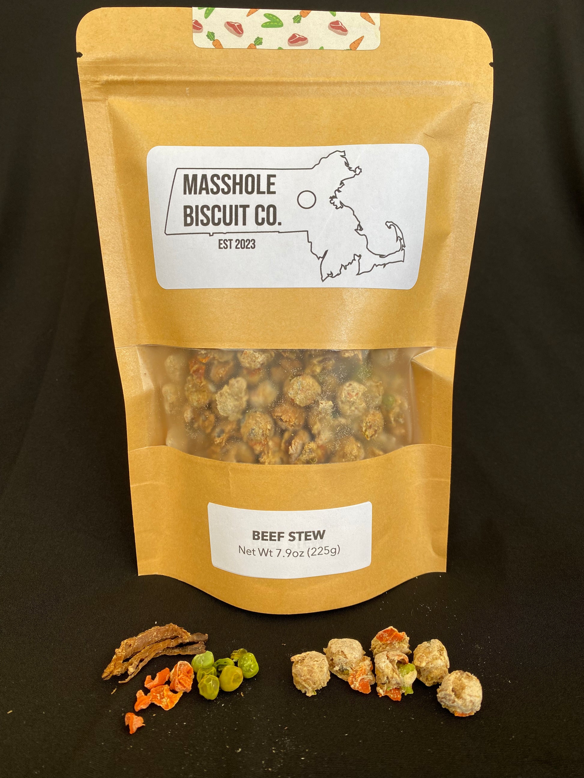 Masshole Biscuit Co all natural dog treat medium training treat bag that features Masshole Biscuit Co training dot-shaped treats. A small pile of these roud training treats are shown next to USDA beef, peas, and carrots, which serve as the chunky style add-ins to the dog treat dough. The Masshole Dog treat package is beige and features the Masshole Biscuit Co logo and the dog treat flavor, beef stew. There is a clear window to see the Massachusetts state-shaped treats.